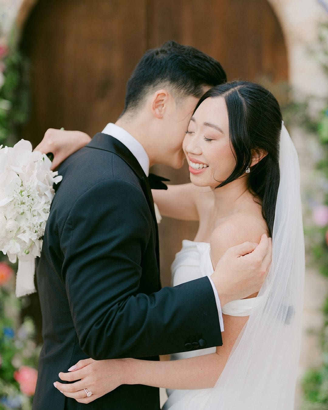 Pareja de recién casados abrazándose, simbolizando el hilo rojo del destino, con la novia sosteniendo un ramo de flores blancas.
