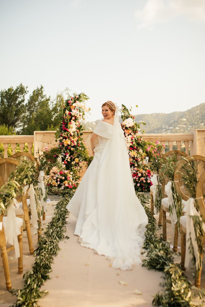 Stephanie walking down an aisle of olive branches and coral blooms at Hotel Cap Vermell, Mallorca