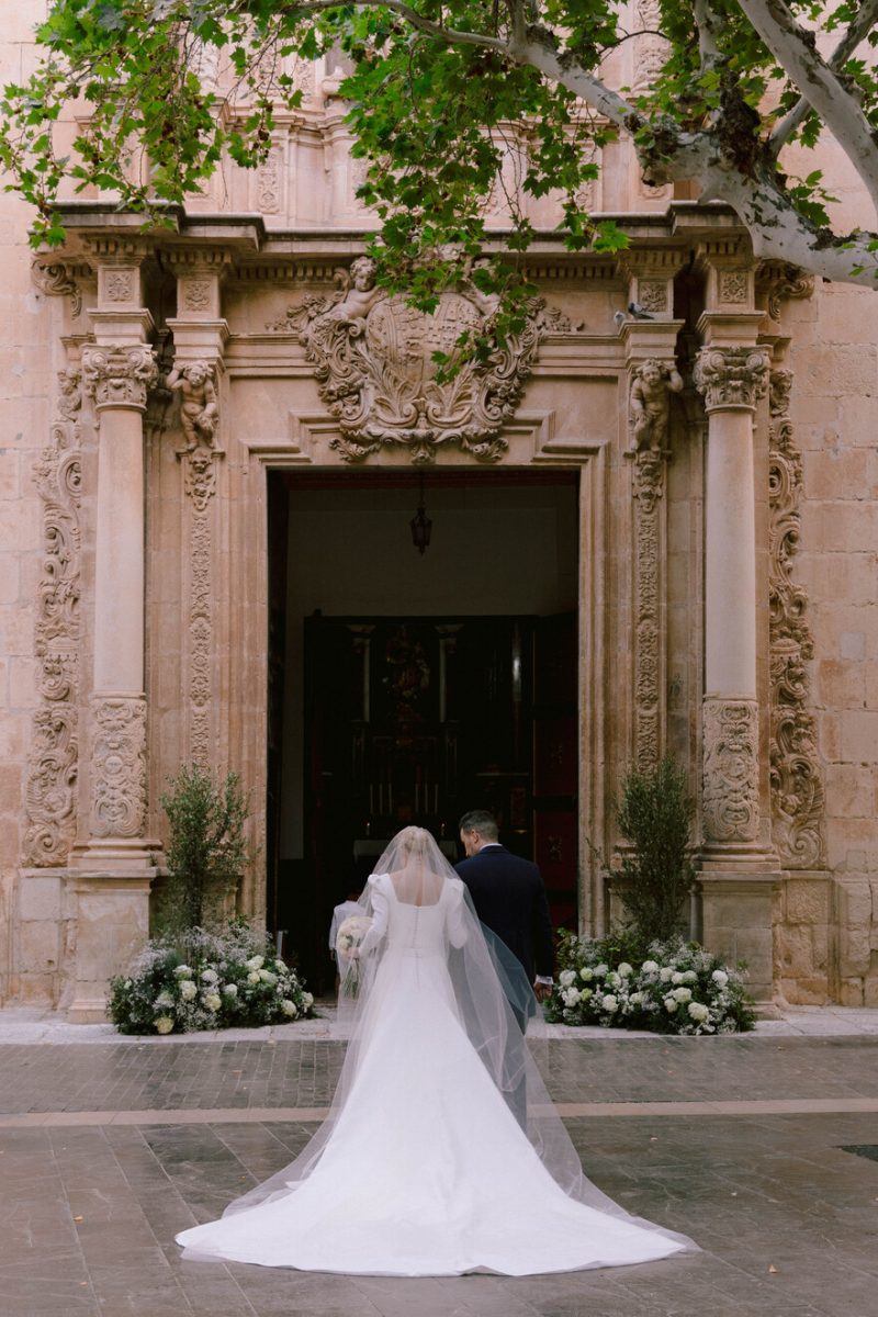 Gema walking in through the baroque stone doorway, her train spread behind her, at her wedding in Alicante